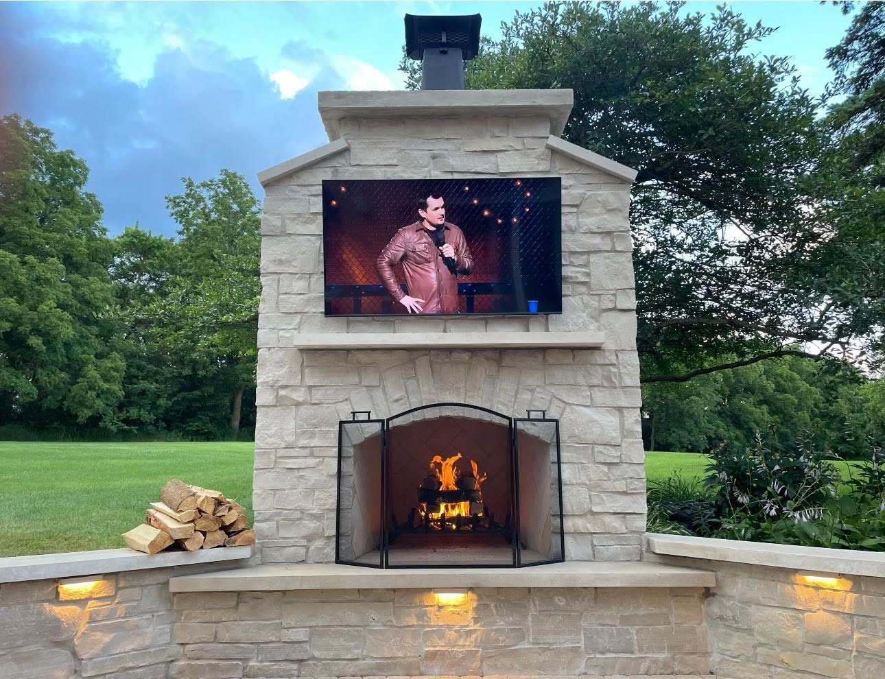 Limestone outdoor fireplace with a wood fire, mounted TV and lit seat walls